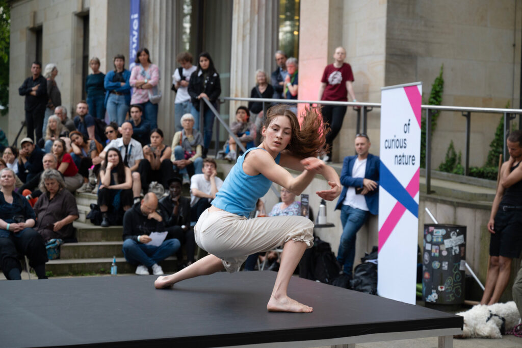 Eine junge Tänzerin macht einen Auftritt auf einer Bühne. Das Publikum sitzt auf Stufen und schaut zu.